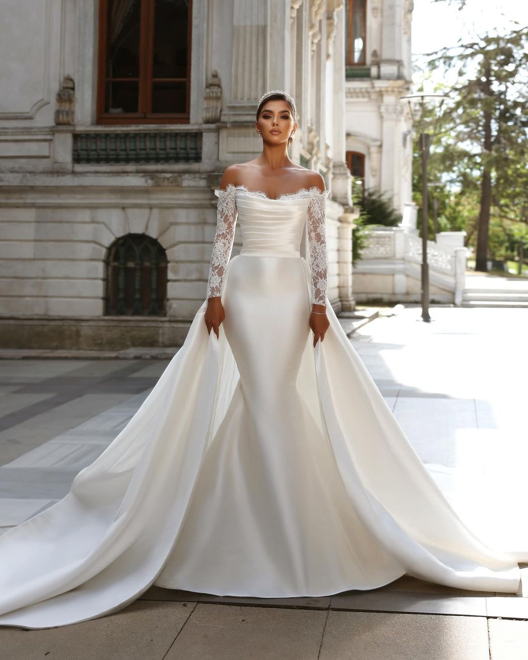 Vestido de novia largo blanco con escote sirena, mangas largas y cola desmontable.